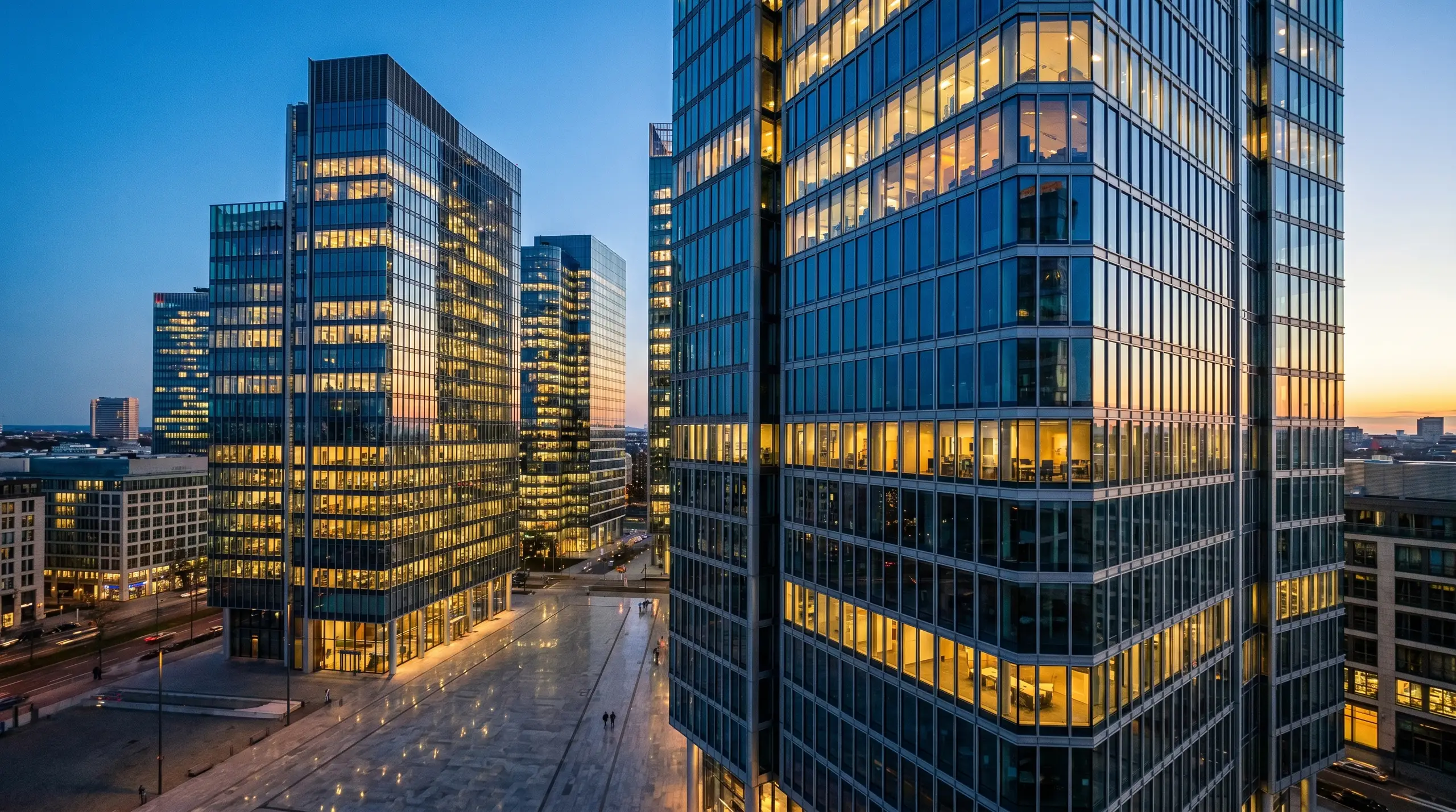 A cluster of tall office buildings with blue-tinted glass and lit windows at dusk. In the foreground, a large building curves along its facade with warm interior lights reflecting in the glass, while a street with parked cars and pedestrians runs in­