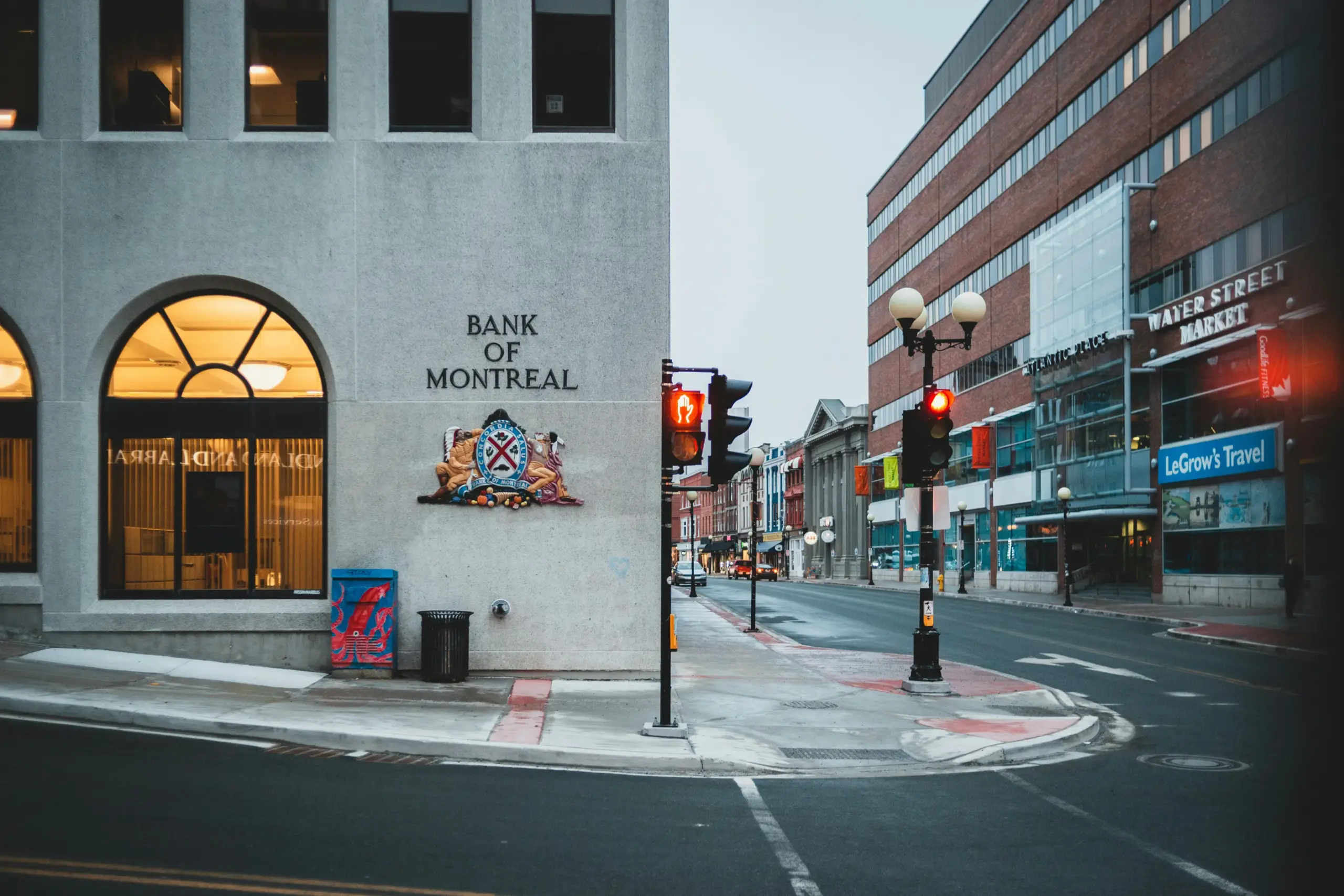 The Bank of Montreal building facade with the words "BANK OF MONTREAL" and a crest above the doorway, facing a city street with a red traffic light and storefront signs including "WATER STREET MARKET" and "LeGrow's Travel".