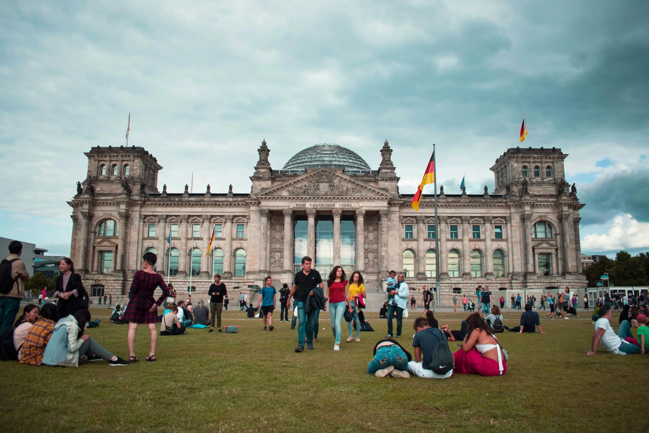 The historic Reichstag building in Berlin stands behind a grassy lawn filled with people relaxing and walking. German flags fly from poles, and the inscription above the entrance reads "DEM DEUTSCHEN VOLKE" in front of the glass dome.
