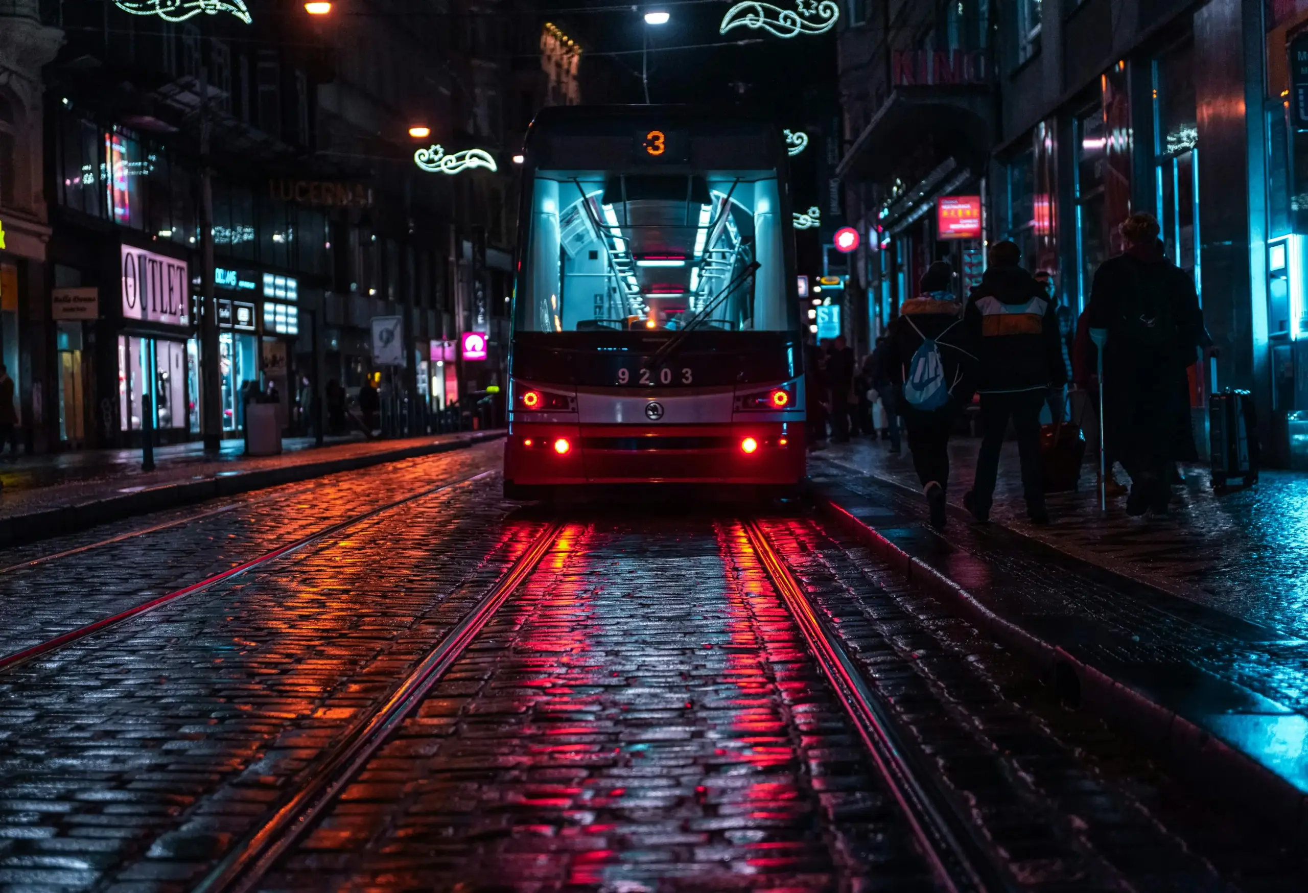 A red futuristic tram with the number 9203 drives toward the camera on shiny wet street tracks at night, with neon-lit storefronts and pedestrians walking along both sides.