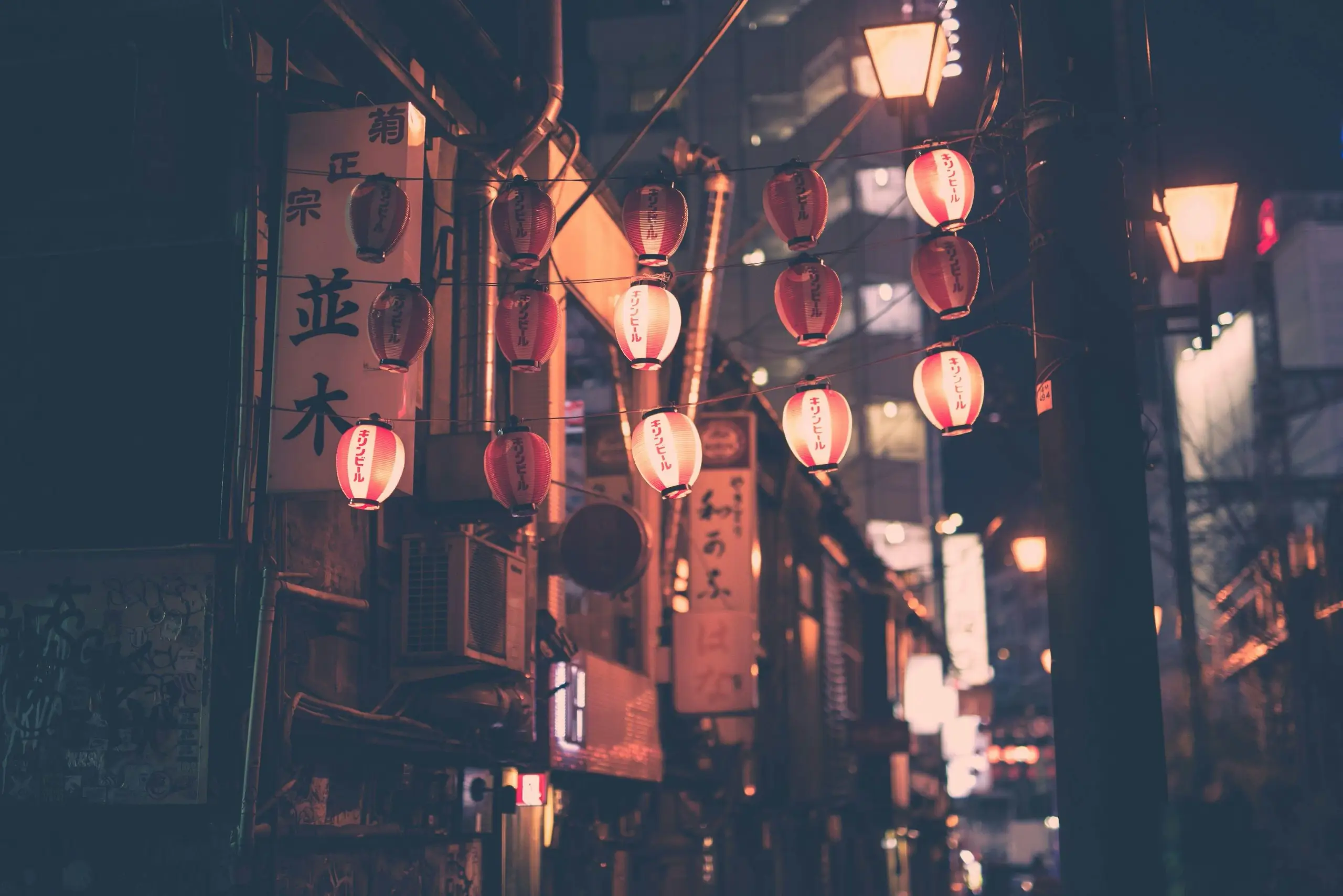 Dozens of red-and-white paper lanterns with Japanese characters hang above a narrow city street at night, with warm streetlights and blurred shop signs in the background.