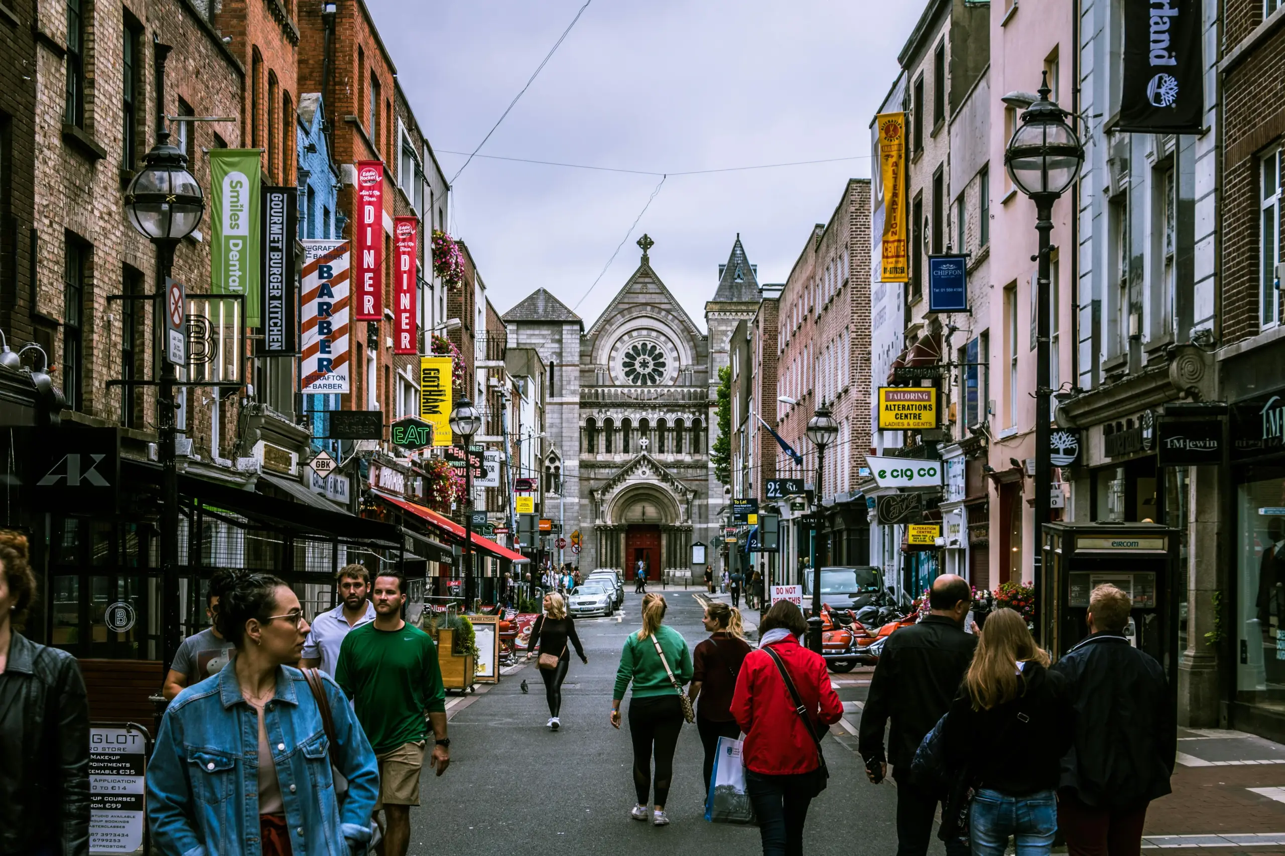 A busy narrow street lined with shops and colorful vertical banners, pedestrians walking toward the distance, and a large stone church with a circular rose window centered at the end of the road.