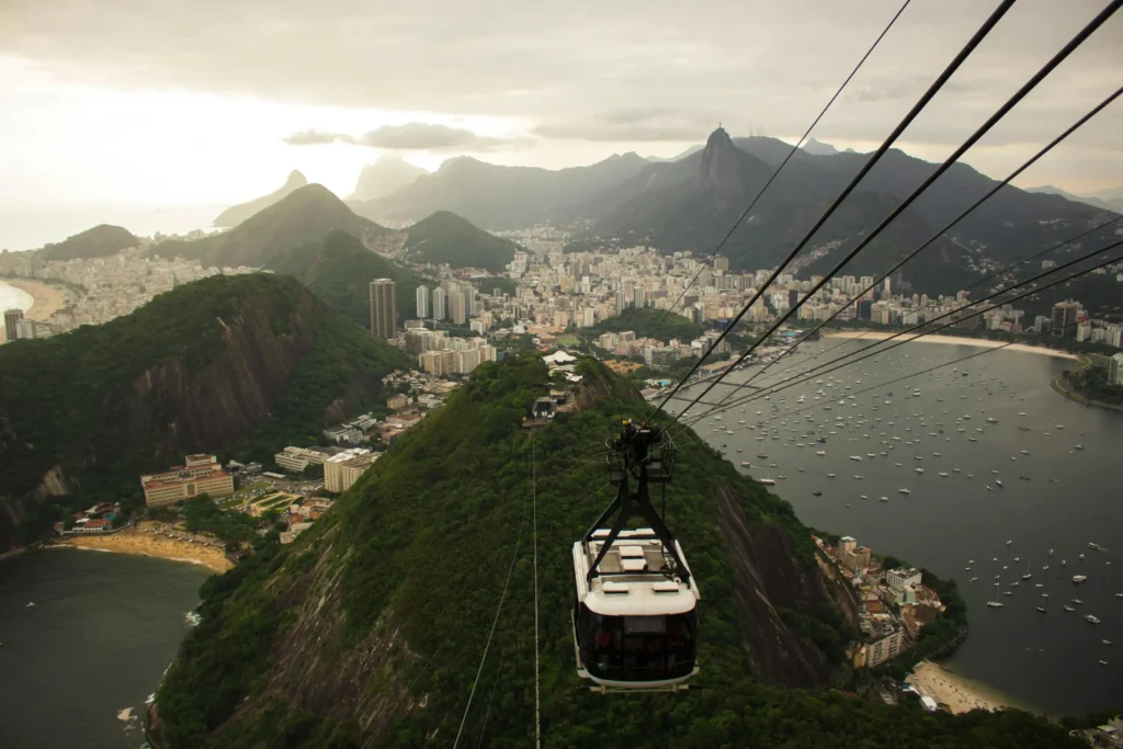 A cable car gondola hangs over a coastal city with a large harbor full of small white boats, surrounded by dark green mountains at sunrise, with cables stretching diagonally across the image.