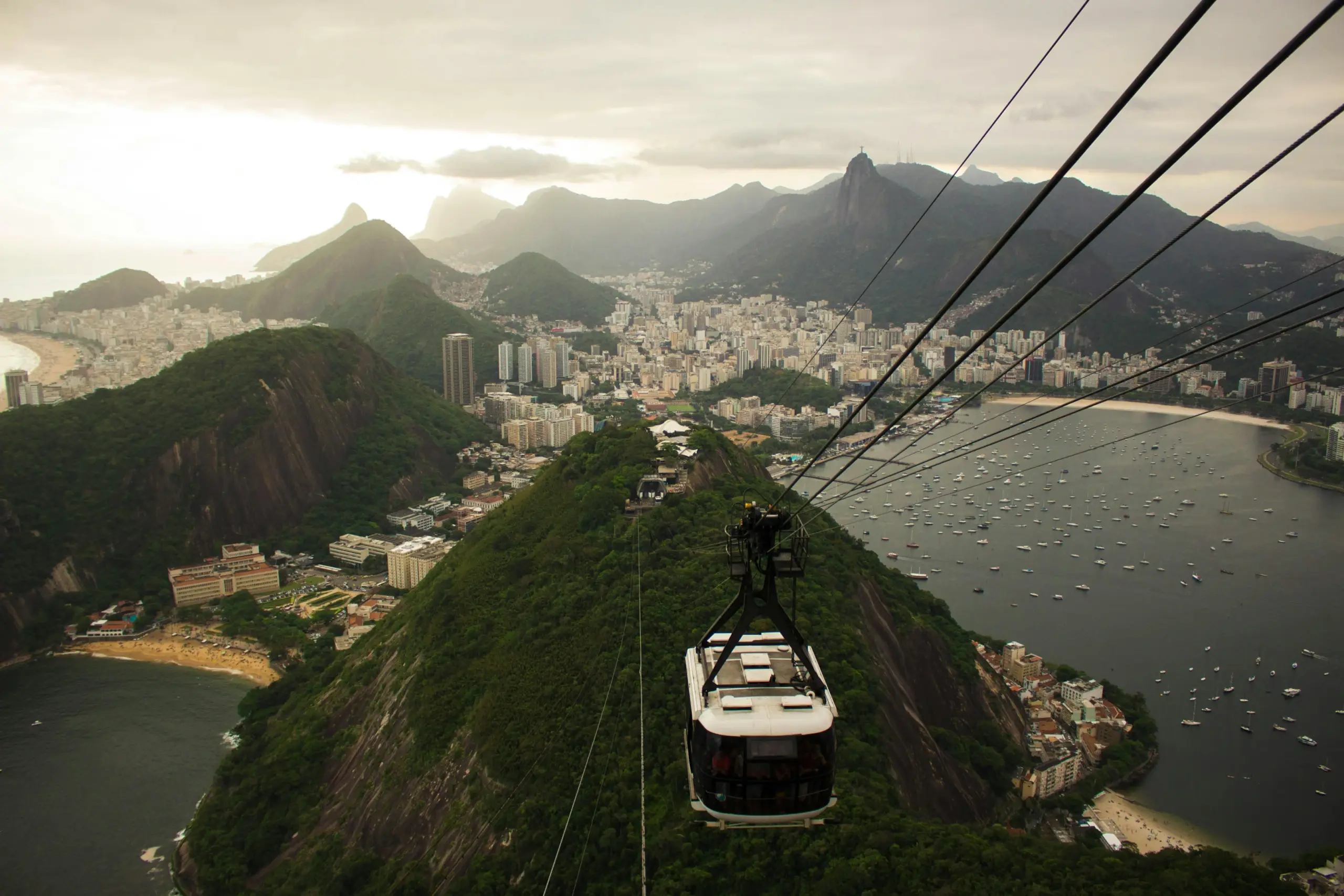 A cable car gondola hangs over a coastal city with a large harbor full of small white boats, surrounded by dark green mountains at sunrise, with cables stretching diagonally across the image.