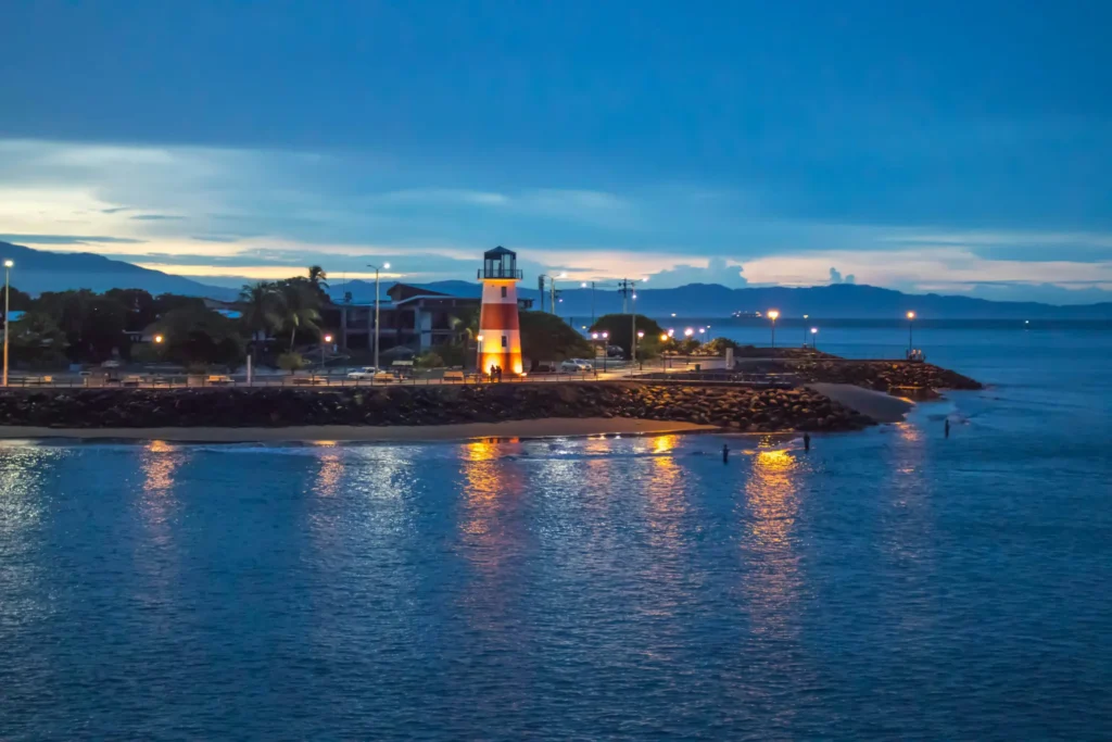 A lit lighthouse stands on a rocky shoreline at twilight, with calm blue water in the foreground, streetlights along the coast, and distant hills under a deep blue sky.