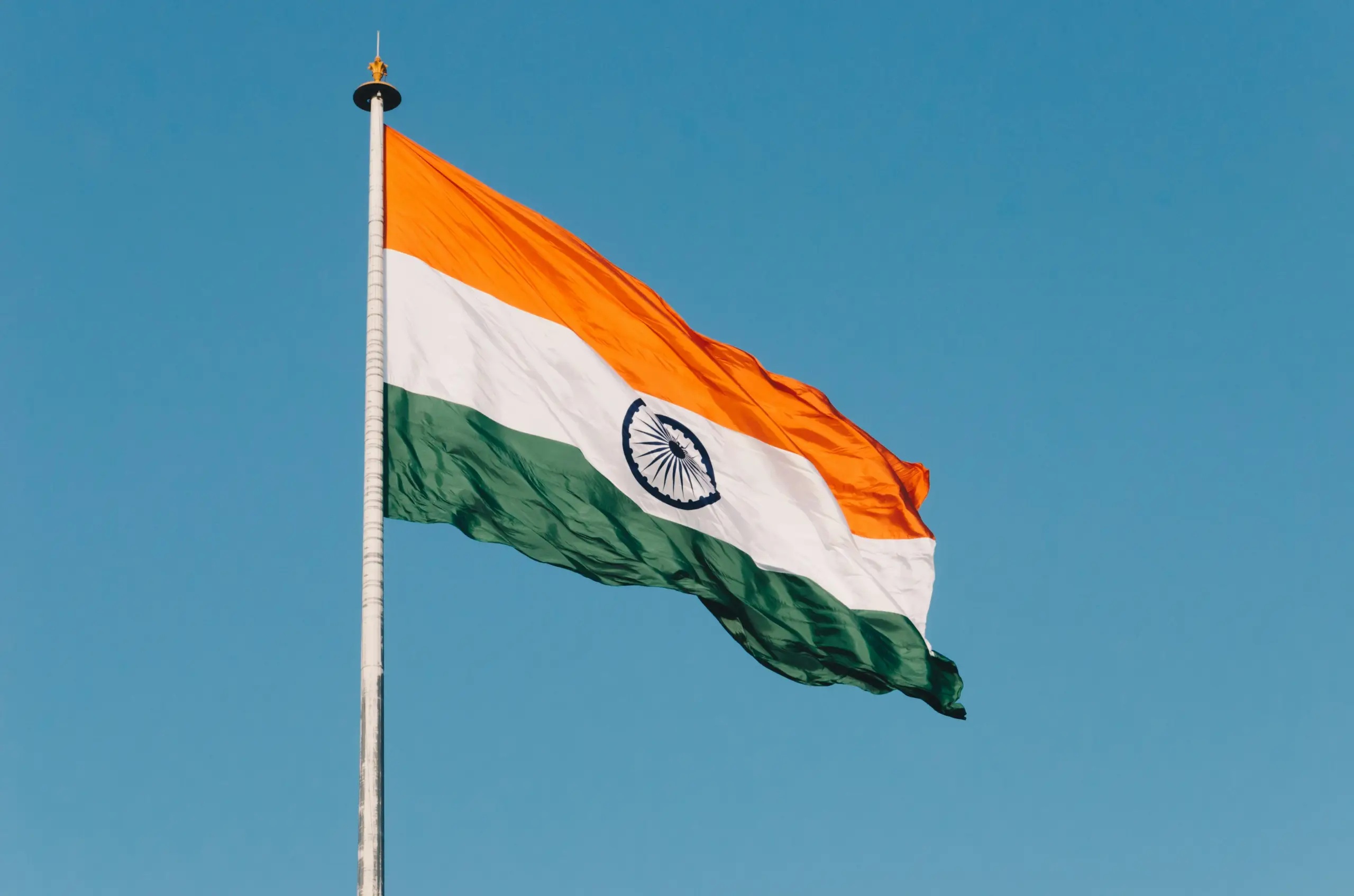The Indian national flag waves on a flagpole against a clear blue sky, showing saffron, white, and green bands with the navy blue Ashoka Chakra in the center.