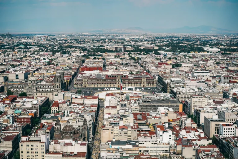 Wide aerial view over a dense city of low-rise buildings and streets, with a long main avenue stretching into the distance and faint hills on the horizon under a blue sky.