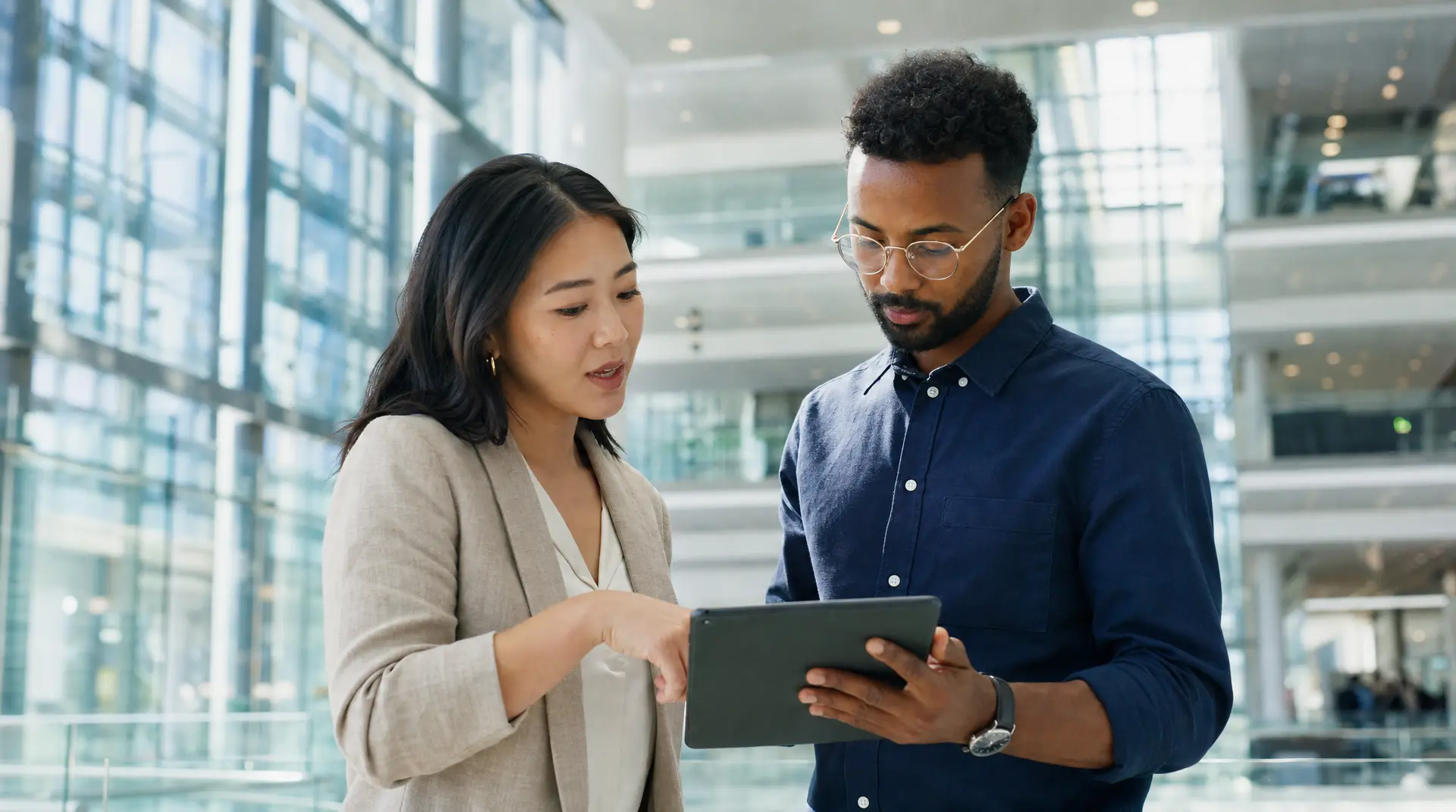 Two professionals review information on a tablet in a modern glass office atrium, supported by compliance ai tools.