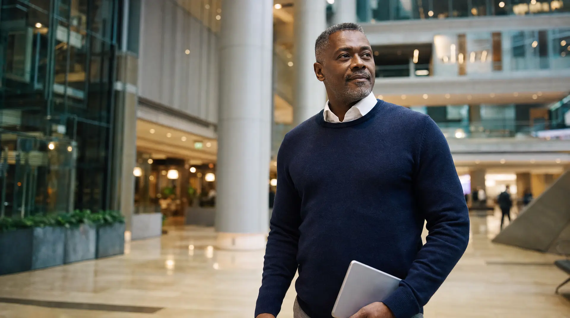 A business professional standing in a modern office lobby holds a tablet while preparing for corporate compliance work with ai tools.