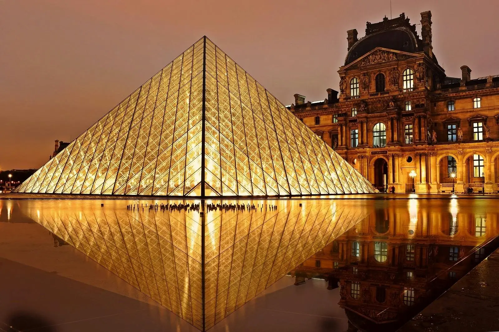 The illuminated Louvre Pyramid rises beside historic museum buildings at dusk, with the scene reflected in the water basin below. The glass pyramid's triangular panels glow yellow under warm lights.