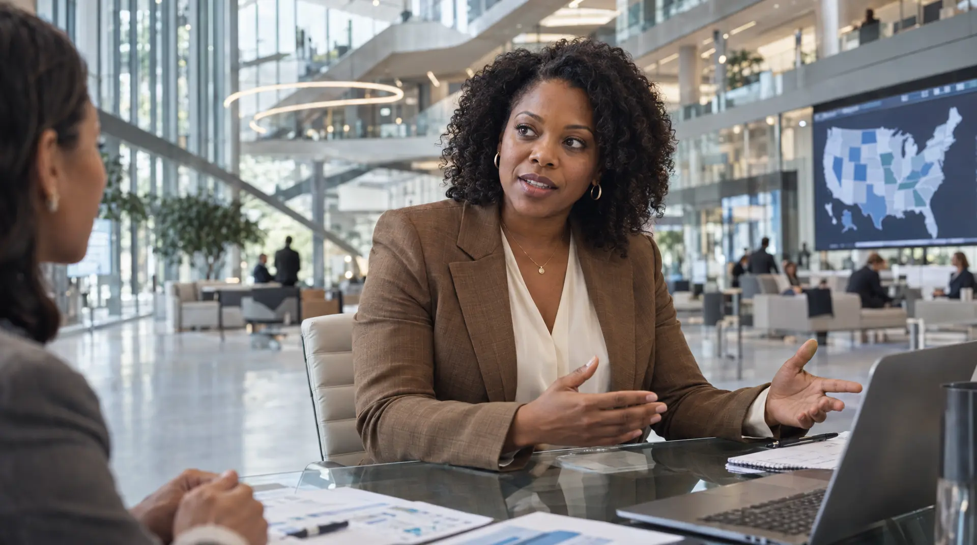 Curly-haired professional in a glass office meeting room discusses documents while a compliance calendar is referenced on a laptop.