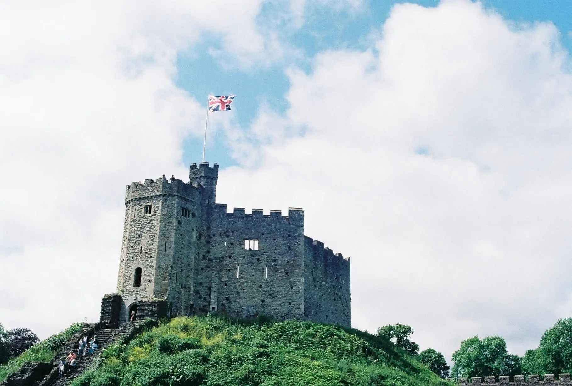 A tall stone castle tower with battlements and narrow windows rises above green trees and shrubs, while a flagpole on the roof flies a Union Jack (red, white, and blue) against a bright blue sky with scattered white clouds.